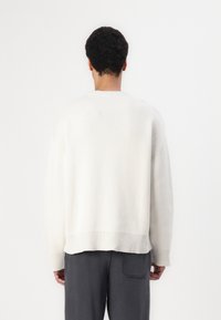 Pulover cu guler rotund în crem deschis, textură tricotată, croi lejer, margini și manșete cu nervuri, asortat cu pantaloni gri închis, văzut din spate.