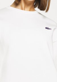Vit sweatshirt i bomull med ribbad rundhals. Har en liten, broderad logotyp på vänster bröst.