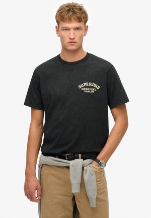 Black cotton t-shirt featuring a faded print "Superdry Harajuku Tokyo Japan" in cream. Short sleeves and round neck, paired with tan trousers.