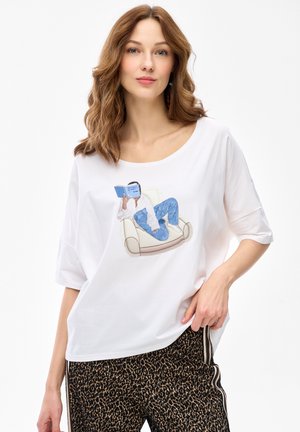 Kobieta o falowanych brązowych włosach, ubrana w białą koszulę z grafiką osoby czytającej na krześle oraz w spodnie w panterkę z bocznymi lampasami.