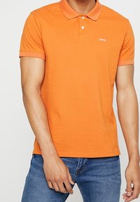 Polo arancione a maniche corte in cotone, con colletto classico e un logo discreto sul petto, abbinato a jeans in denim.