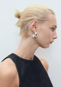Stříbrné kovové kruhové náušnice s hladkým, zaobleným designem. Nosené na modelce s krátkými blond vlasy, která má na sobě tmavý top bez rukávů.