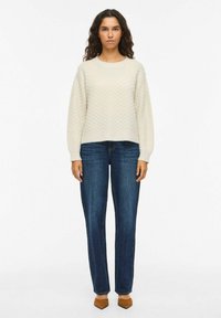 Pull à texture crème avec un col rond, associé à un jean droit foncé et des chaussures à talons marron. Le mannequin se tient devant un fond uni.