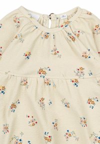 Rochie cu model floral în crem, cu pliuri la gât. Prezintă flori mici, multicolore și un sistem de închidere cu buton la spate.