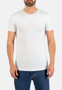 Mann, der ein schlichtes, weißes, enges T-Shirt mit kurzen Ärmeln und bluejeans trägt, steht vor einem weißen Hintergrund.