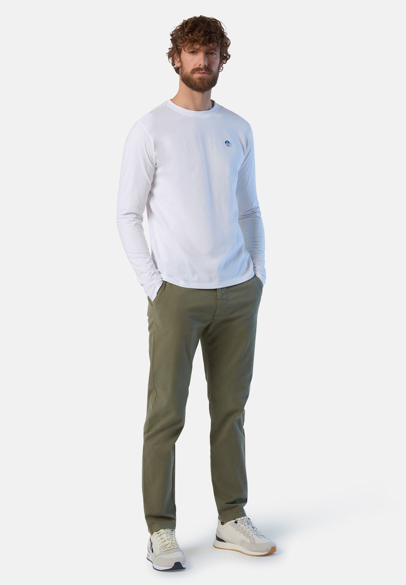 Camicia bianca a maniche lunghe con un piccolo logo blu, pantaloni verde oliva e sneaker chiare con accenti grigi e beige. Posizione in piedi.