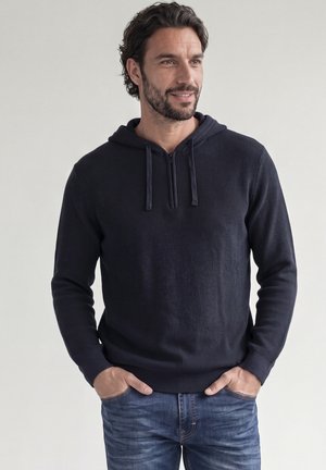 Mann mit dunklem Haar und Bart, der einen marineblauen Kapuzenpullover und blaue Jeans trägt, die Hände in den Taschen, schaut leicht nach rechts und lächelt leicht.
