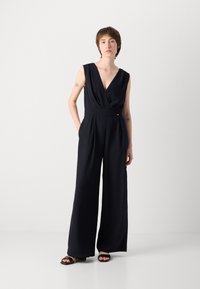 Mørkeblå uten ermer jumpsuit med dyp V-hals, vide ben og en tettsittende liv. Stoffet er glatt og flytende, med diskrete sidelommer.
