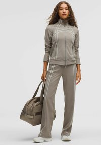 Szürke sportos melegítő összeállítás fehér díszítő varrásokkal, cipzáras dzsekivel és gumírozott derekú nadrággal. A modellen egy hozzá illő sporttáska is látható.