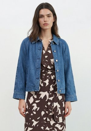 Vrouw met bruin haar tot op de schouders, die een blauwe spijkerjas draagt over een bruin-witte jurk met een abstract patroon en een tailleband.