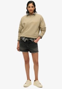 Sudadera beige con puños acanalados, sobre una camisa de cuadros. Pantalones cortos de mezclilla negros con dobladillo deshilachado. Zapatillas crema que completan el conjunto.