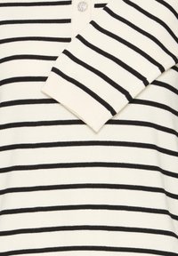 Haut en maille blanc à manches longues avec des rayures noires horizontales, poignets côtelés et un bouton orné de bijoux près du col.
