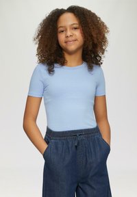 Top corto de manga corta de color azul claro con ribetes, combinado con pantalones de mezclilla anchos de color oscuro que tienen una cinturilla elástica y cordón, con las manos en los bolsillos.