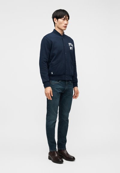 Άνδρας που στέκεται, φοράει ένα σκούρο μπλε σακάκι με κουμπιά και το κείμενο "Polo NY", μπλε τζιν και σκούρα καφέ δερμάτινα μποτάκια σε απλό λευκό φόντο.