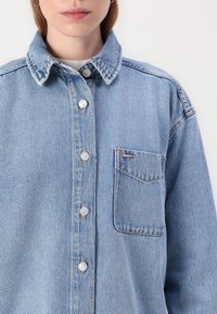 Camicia in denim azzurro chiaro con colletto classico, bottoni frontali e taschino sul petto con dettagli di cucitura discreti.