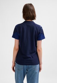 Mörkblå pikétröja med korta ärmar, med en slät textur och kontrasterande mörkblå axeldetaljer. Avslappnad passform, kombinerad med ljusblå jeans.