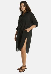La modella sta in piedi con sicurezza indossando un vestito camicia nero con maniche arrotolate e spacchi laterali, abbinato a sandali marroni, mentre sorride dolcemente.