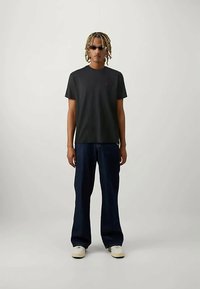 Jauns cilvēks ar blondiem dreads, tērpies melnā T-kreklā, tumšās platās biksēs, baltos sporta apavos un šaurās melnās saulesbrillēs, stāv uz neitrālas fona.