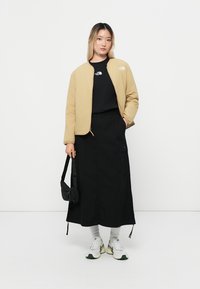 Veste bomber marron clair sur un sweat noir, associée à une longue jupe noire et des baskets multicolores. Elle tient un petit sac noir.