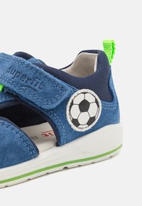 Blå semsket sandal med sjøgrønne tekstildetaljer, grønn stropp og et fotballmerke. Har en hvit såle med en grønn linje.