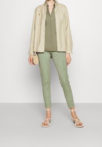 Camicia beige con bottoni, top verde oliva, pantaloni aderenti verde chiaro e sandali crema con cinturini. Borsa a tracolla minimalista.