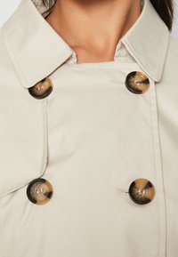 Trench-coat beige présentant un design à double boutonnage, de grands boutons ronds marron, et un col classique avec une texture lisse et brillante.