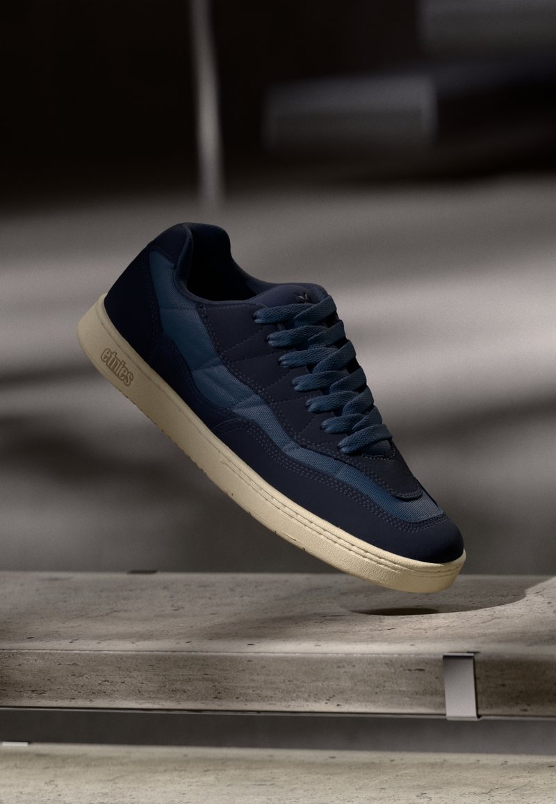 Sneaker blu navy con suola beige e logo di lato, sospesa sopra una superficie di pietra in un'ambientazione interna poco illuminata.