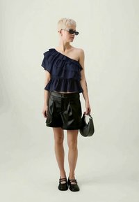 Top azul-marinho com folhos de um ombro, calções pretos em pele sintética e bolsa preta; modelo usa óculos de sol e sapatos pretos.