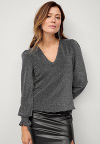 Grå, langærmet indpakningsbluse med V-hals og bølgede manchetter, lavet af struktureret, metallisk stof. Parret med en sort, kunstlæder nederdel.