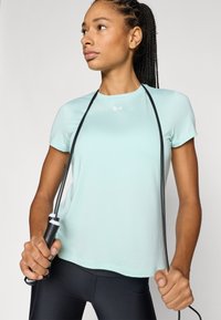 Ανοιχτό μπλε αθλητικό μπλουζάκι με κοντά μανίκια, συνδυασμένο με μαύρα κολάν. Ένα σχοινάκι για άλματα κρατιέται και από τα δύο χέρια, δείχνοντας τη λεπτομέρεια της λαβής.