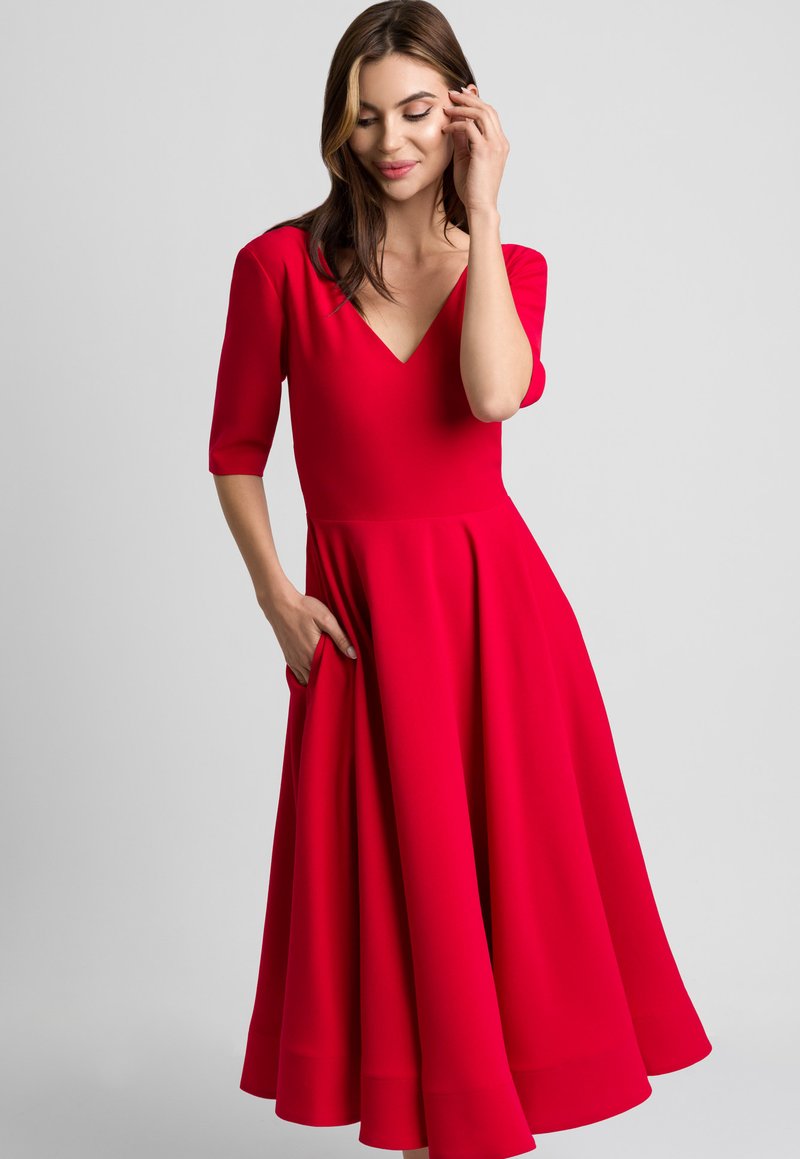 Kobieta w czerwonej sukience z dekoltem w serek i rękawami do łokcia, z rozkloszowanym dołem, z jedną ręką w kieszeni, stojąca na tle jasnoszarym.