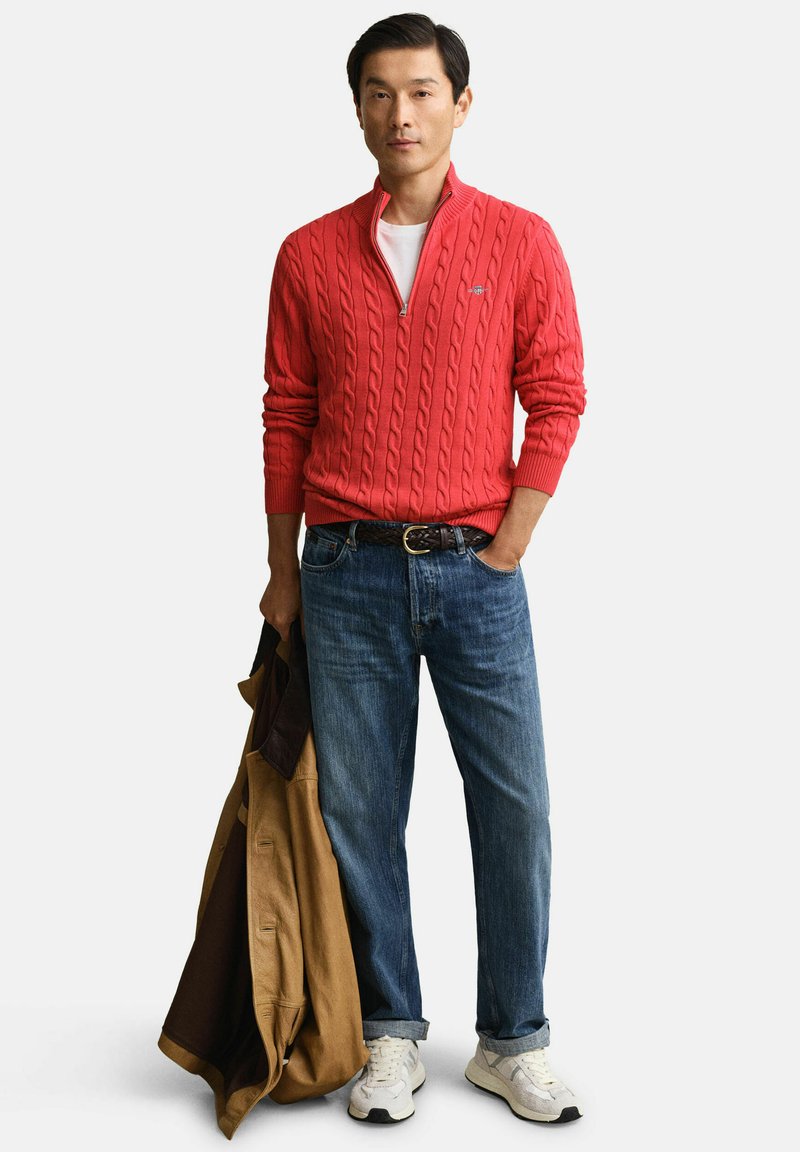 Uomo che indossa un maglione lavorato a trecce color corallo, jeans blu, sneakers bianche, tiene in mano una giacca color cuoio con la mano sinistra, in piedi davanti a uno sfondo neutro.