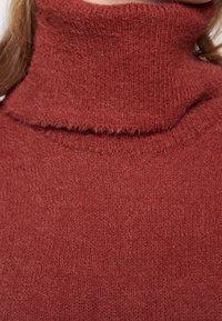 Červený pletený rolák s texturovaným, měkkým povrchem. Má obrácený výstřih a volný střih, což zdůrazňuje teplo a pohodlí.