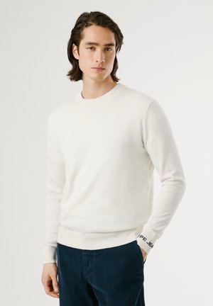 Suéter de cuello redondo de color beige claro en una tela suave, con mangas largas y puños acanalados, que presenta un logotipo negro contrastante en la muñeca derecha.