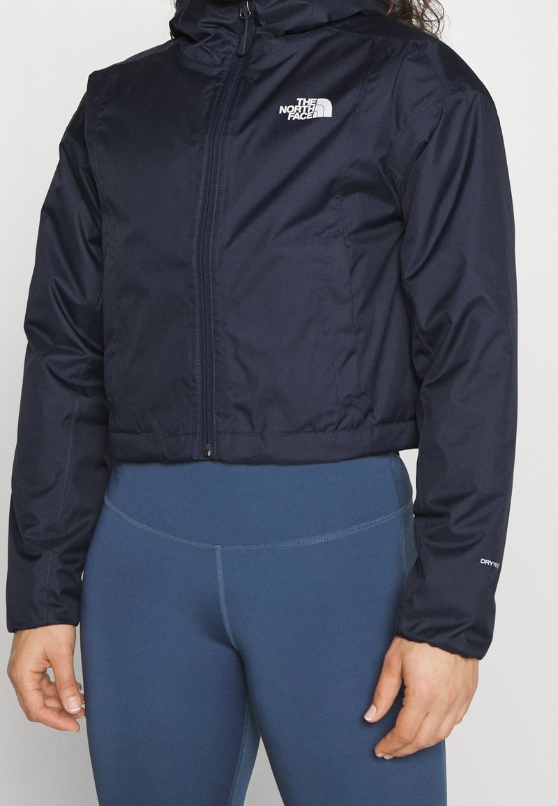 Navy blue cropped jacket with a hood, zip closure, and a white logo on the left chest. Features elastic cuffs and a smooth, textured exterior.