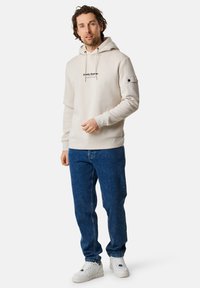 Felpa beige in cotone con logo frontale, cappuccio con cordino e polsini a costine, abbinata a jeans in denim blu e sneakers bianche.