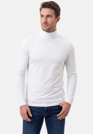 Mann trägt ein weißes langärmeliges Rollkragenshirt und blaue Jeans, steht mit einer Hand in der Tasche und blickt zur Seite.