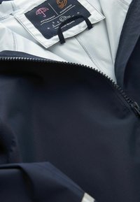 Vodootporna mornarsko plava jakna iznutra prikazuje svijetlu sivu podlogu, patentni zatvarač, oznaku za njegu s ikonama, te slojevite šavove za poboljšanu izdržljivost.