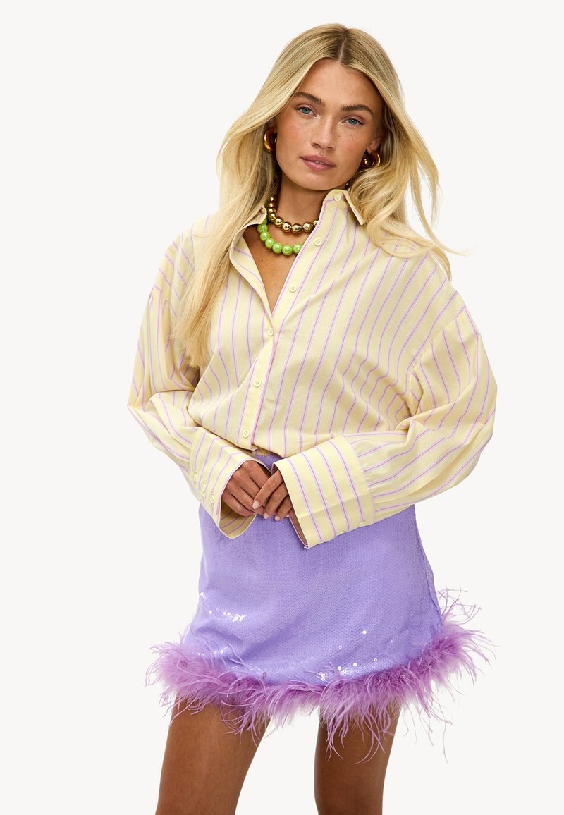 Chemise surdimensionnée rayée jaune et rose associée à une mini-jupe en sequins lilas avec une bordure en plumes roses duveteuses. Collier en perles multicolores.