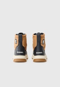 Botas de tobillo marrones y negras con parte superior acolchada, logo de SOREL prominente, suelas de goma texturizadas y tiradores en la parte posterior para ponerlas y quitarlas fácilmente.