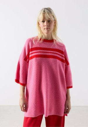 Young woman wearing an oversized pink sweater with red stripes and red pants, standing against a plain white background.