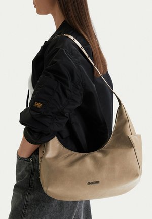 Mujer con chaqueta bomber negra y jeans grises, llevando un bolso de cuero beige con tachuelas en la correa.