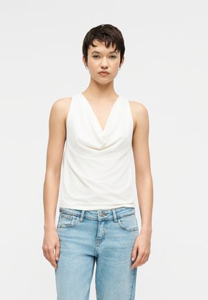 Νεαρός με κοντά σκούρα μαλλιά, που φοράει λευκό αμάνικο τοπ με πτυχώσεις και ανοιχτόχρωμο τζιν, στέκεται μπροστά σε απλό φόντο.