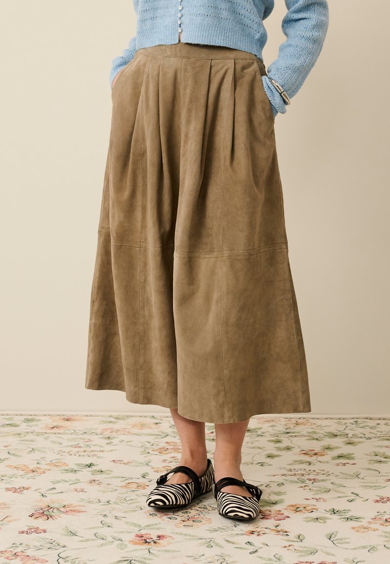 Beige suède geplooide rok met een losse pasvorm, voorzien van zijzakken en een gladde textuur. Gecombineerd met zwart-wit gestreepte platte schoenen.