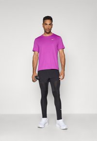 Lilla kortærmet træningsshirt med et lille hvidt logo, sorte tætsiddende shorts, sorte leggings og hvide sneakers. Holder en vandflaske.