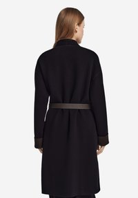 Langer schwarzer Mantel mit tailliertem Gürtel, ausgestattet mit hohem Kragen und kontrastierenden braunen Manschetten. Weiche Textur mit geradem Schnitt.