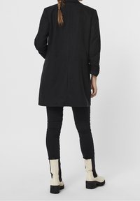 Musta pitkä takki, jossa on mittatilaustyönä tehty muotoilu, sivuleikkaukset ja käännetyt hihat, yhdistettynä mustiin skinny-farkkuihin ja vaalean kremankkoihin nilkkabootseihin.