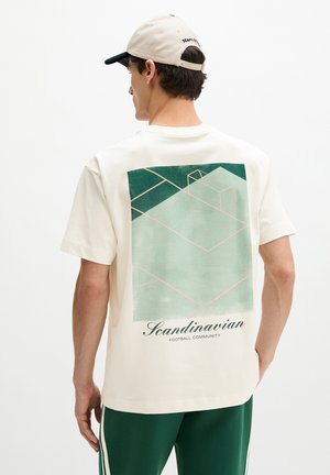 Junger Mann trägt ein weißes T-Shirt mit einem grünen geometrischen Fußballfeld-Design und dem Text "Scandinavian Football Community" auf dem Rücken.