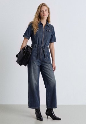 Donna che indossa una tuta di jeans blu con maniche corte, tiene in mano una borsa nera e indossa stivaletti neri con tacco.
