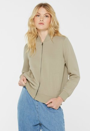 Blonde woman wearing beige zip-up jacket and loose blue jeans, posing with one hand in pocket against plain background.